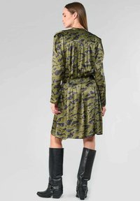 Grünes gemustertes Kleid mit langen Ärmeln, tailliertem Bund und entspannter Passform. Schwarze kniehohe Stiefel mit Schnallenakzenten. Glatte Stofftextur.