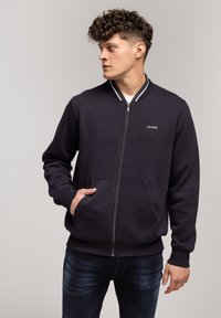 Granatowy sweatshirt na zamek z prążkowanym kołnierzem, przednimi kieszeniami i małym logo na piersi. Wykonany z miękkiego, teksturowanego materiału.
