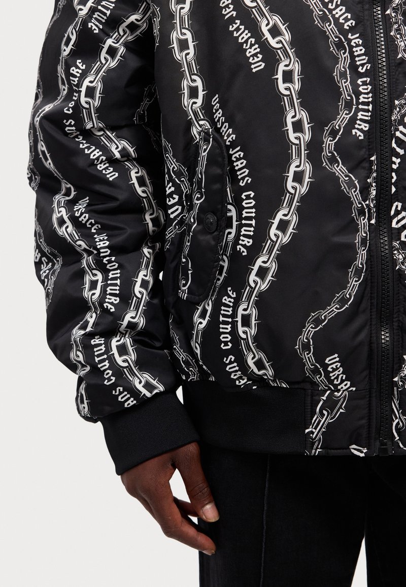 Giacca bomber nera con motivo a catena e testo bianco, con una finitura lucida, polsini a coste e chiusura con cerniera. Presenta anche una tasca laterale.