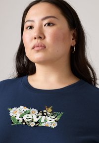 Μπλε ναυτικό μπλουζάκι με λουλουδάτο σχέδιο "Levi's" που απεικονίζει μαργαρίτες και μια πεταλούδα. Σχέδιο με λαιμόκοψη και κοντά μανίκια με απαλή υφή.