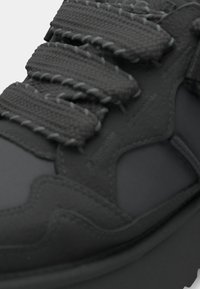 Svart sneaker med texturerat material, en lager-på-lager design och tjocka vävda remmar. Inkluderar en slät gummisula och utskurna detaljer.