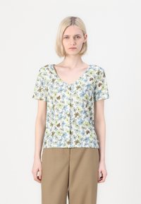 Una donna con capelli biondi corti che indossa una maglietta a maniche corte con bottoni, caratterizzata da una stampa floreale blu, verde e marrone, e pantaloni marrone chiaro.