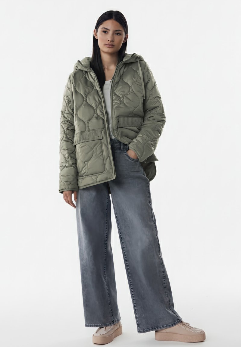 Giacca trapuntata verde oliva con cappuccio, due tasche frontali, orlo a coste, abbinata a jeans grigi a gamba larga e scarpe beige con plateau.