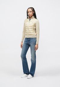 Μπεζ φουσκωτό μπουφάν με φερμουάρ σε συνδυασμό με κρέμ μανίκια, συνδυασμένο με ανοιχτό μπλε καμπάνα τζιν και λευκά αθλητικά παπούτσια.