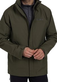 En person i en olivgrön jacka med ett dragkedjeskyddat innerlager justerar frontstängningen och visar upp en avslappnad utomhusstil.