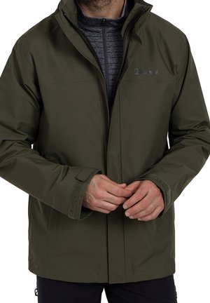 Une personne portant une veste vert olive avec une couche intérieure zippée ajuste la fermeture à l'avant, mettant en avant un style décontracté de plein air.
