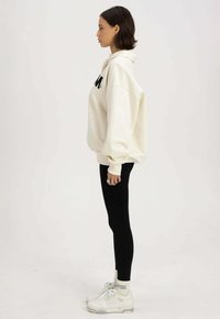 Crème oversized hoodie met zwart logo, gecombineerd met zwarte leggings en witte sneakers met grijze accenten en textuurdetails.