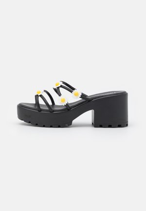 Sandales à plateforme noires avec trois fines lanières ornées de marguerites blanches et de centres jaunes. Semelle texturée pour une meilleure adhérence. Design à bout rond.