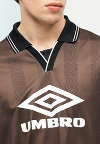 Mlada oseba, oblečena v rjavo športno majico Umbro s črnim ovratnikom, na katerem so bele dvojne črte in geometrijski vzorec.