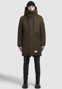 Cămașă parka de culoare verde măslinie cu guler înalt, glugă ajustabilă, buzunare frontale și etichetă de marcă; asortată cu blugi negri și bocanci.