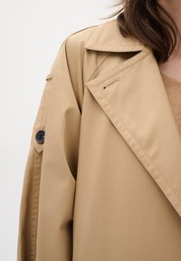 Light beige trench coat made of smooth, textured fabric. Features a classic collar, shoulder button detail, and visible stitching.