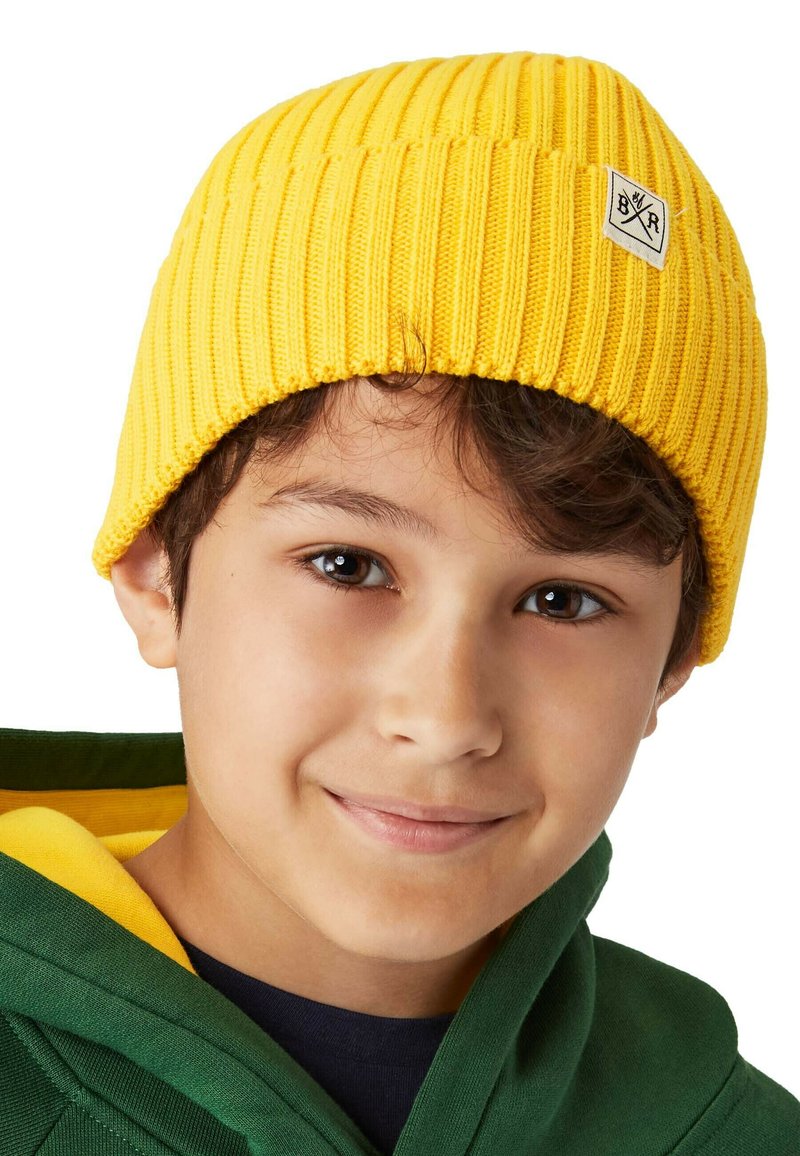 Gul stickad mössa med ribbat mönster, prydd med en liten vit etikett. Subjektet bär en grön hoodie under mössan.