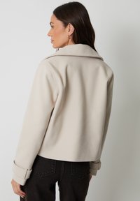 Beige uldblandingsjakke med klapskrave og rullede ærmer, med et cropped design. Glat tekstur og minimal detaljer.
