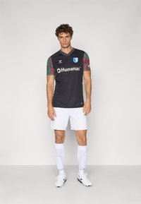 Black short-sleeve football jersey with patterned green sleeves, featuring white logo and text, worn with white shorts and knee-high socks.