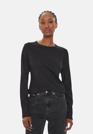 Donna con i capelli intrecciati che indossa una maglia nera a maniche lunghe e jeans scuri a vita alta, in piedi davanti a uno sfondo chiaro e uniforme.