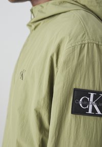 Ljusgrön huvjacka i texturerad tyg, med en svart lappar med vit logga på höger ärm och minimalistisk design.