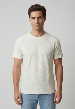 Uomo con capelli corti e castani, che indossa una maglietta bianca a maniche corte texturizzata e jeans azzurri, in piedi davanti a uno sfondo semplice.