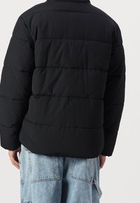 Chaqueta negra acolchada con diseño acolchado, con cuello alto y mangas largas. Combinada con vaqueros cargo azul claro.