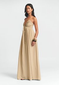 Maxi-mekko beige-värissä, jossa on syvä V-mallinen kaula-aukko, rypytetty bodice, säädettävä nauha ja valuva kangasrakennus. Koristeltu pinotuilla rannekoruilla.