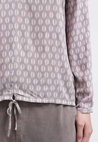 Chemise à manches longues gris clair avec un motif géométrique en blanc et rose pastel, dotée d'un cordon de serrage à l'ourlet et d'une texture douce.