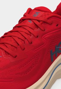 Κόκκινο αθλητικό παπούτσι με λεπτομερείς ραφές, κορδόνια στο ίδιο κόκκινο χρώμα και μπεζ σόλα με μπλε και γκρι λογότυπα.