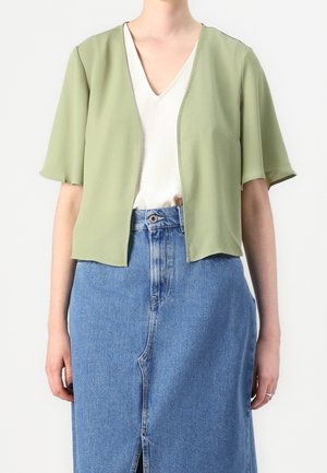 Femme portant un cardigan vert clair à manches courtes et ouvert sur le devant, un top blanc col en V et une jupe en jean bleue taille haute avec une fente à l'avant.