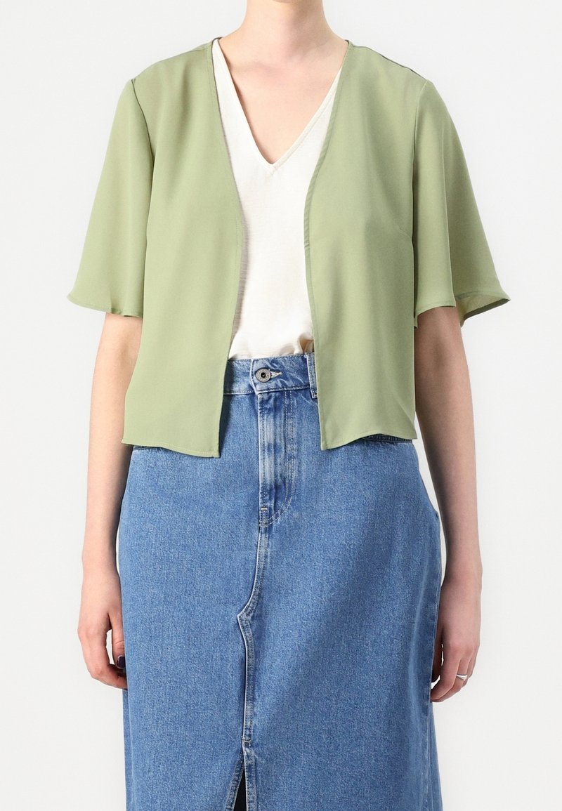 Femme portant un cardigan vert clair à manches courtes et ouvert sur le devant, un top blanc col en V et une jupe en jean bleue taille haute avec une fente à l'avant.