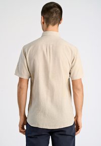 Camisa de manga corta de color beige claro, hecha de tela texturizada, con un cuello estándar y un detalle de pliegue en la parte posterior, combinada con pantalones azul marino.