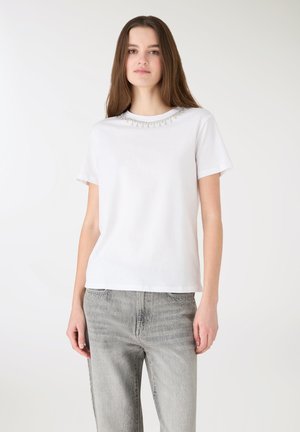 Biały bawełniany t-shirt z krótkimi rękawami, ozdobiony koralikami wokół dekoltu, o luźnym kroju. W połączeniu z jasnoszarymi dżinsami.