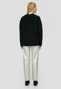 Übergroßer schwarzer Strickpullover mit V-Ausschnitt und ribbeliger Textur, kombiniert mit glänzenden, silbernen weiten Hosen und schwarzen Plateauschuhen.