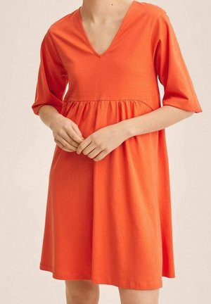 Žena v volných, do kolen sahajících oranžových šatech s výstřihem do V a tříčtvrtečními rukávy, stojící se sepjatýma rukama před sebou.
