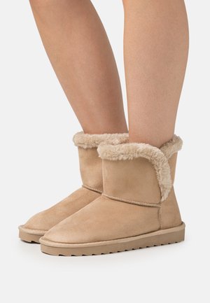 Beige Wildleder-Stiefeletten mit Futter und Schlag aus Kunstfell, flache Gummisohle und genähte Details. Die Stiefel werden auf nackten Beinen getragen.