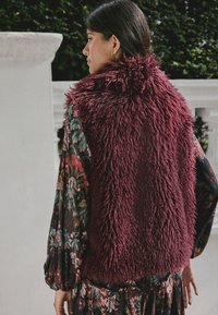 Bordeauxrotes Kunstpelzweste mit hohem Kragen und strukturiertem Design, getragen über einem gemusterten Blumenkleid mit lebendigen Farben und weiten Ärmeln.