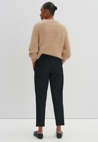 Un pull en beige, duveteux, avec des manches trois-quarts, associé à un pantalon noir à taille élastique et des ballerines noires, vu de dos.