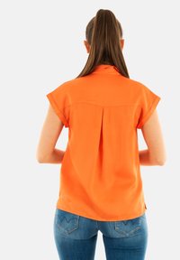 Blusa corta de manga corta en color naranja con un cuello, que presenta un detalle de pliegue en la parte trasera. Combinada con jeans ajustados azules, mostrando un diseño limpio y simple.