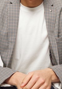 Szary i brązowy marynarka w kratę na gładkiej białej koszulce. Marynarka ma pojedynczy guzik i wzorzystą podszewkę widoczną przy mankietach.