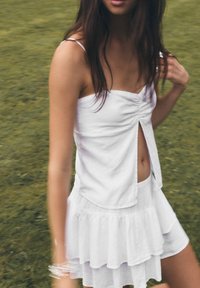 Abito mini bianco a strati con spacco frontale e corpetto arricciato. Presenta spalline sottili e un tessuto leggero e testurizzato.