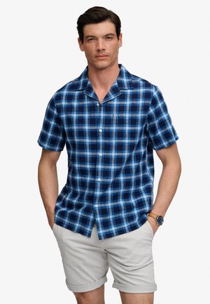 Hombre con camisa de manga corta a cuadros azul y blanca, pantalones cortos enrollados de color beige claro y un reloj de pulsera.