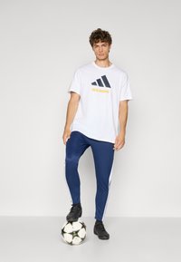 Hvid T-shirt med blå og gul "REAL MADRID" print, parret med marineblå træningsbukser med hvide sidelister. Sort fodbold ved fødderne.