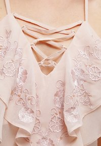Růžový top bez rukávů se rýhovaným předním dílem, zdobený květinovou výšivkou a křížovými ramínky. Lehce průhledná látka s texturovanými detaily.