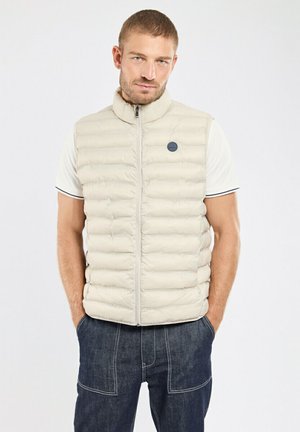 Homme debout les mains dans les poches, portant un gilet matelassé beige sur une chemise blanche à manches courtes et un pantalon bleu foncé, sur fond blanc.