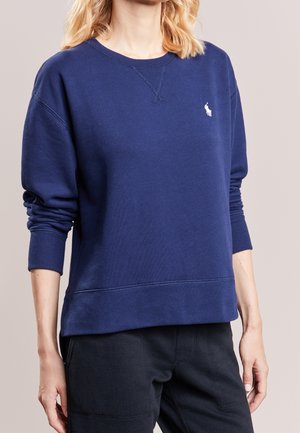 Frau trägt einen marineblauen Pullover mit langen Ärmeln und kleinem weißem Logo auf der linken Brust sowie schwarze Hose.