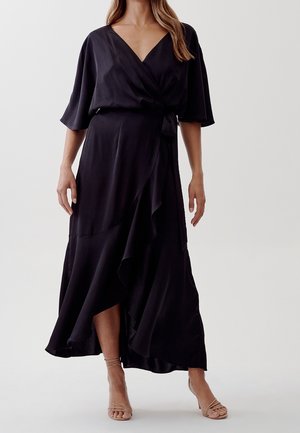 Kobieta ubrana w czarną sukienkę kopertową z rękawami do łokcia i asymetrycznym dołem, w połączeniu z cielistymi sandałkami na obcasie, stoi na jednolitym tle.