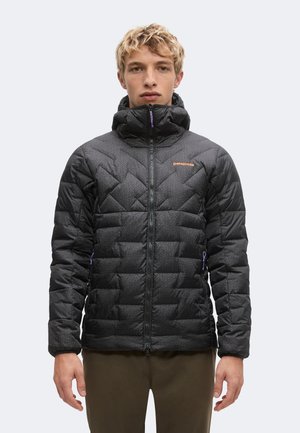 Junge Person mit kurzen blonden Haaren, die eine schwarze, gemusterte Steppjacke mit Frontreißverschluss und hohem Kragen trägt.