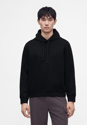 Junger Mann mit schwarzem Kapuzenpullover mit subtil wiederholtem Muster und grauer Hose, stehend vor einem einfarbigen hellen Hintergrund.