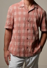 Hombre vestido con una camisa de cuadros texturada de color óxido y de manga corta, combinada con pantalones blancos, de pie frente a un fondo liso.