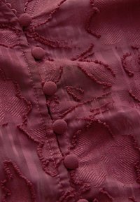 Burgundská látka s texturovanými květinovými vzory a zvýrazněnými akcenty. Šest kulatých knoflíků podél légy, hladká a mírně průsvitná textura.