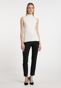 Ærmeløs, højhalset creme top parret med tætsiddende sorte bukser. Modellen står i sorte spidse hæle mod en neutral baggrund.