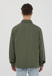 Camicia in cotone verde con polsini abbottonati e colletto a punta, caratterizzata da una texture semplice e un taglio dritto. Nessun logo o motivo visibile.