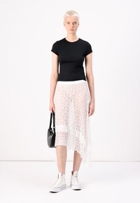 Tricou negru ajustat, fustă albă din dantelă transparentă cu model floral, hem neregulat, purtată cu adidași albi cu guler înalt și o geantă mică neagră.